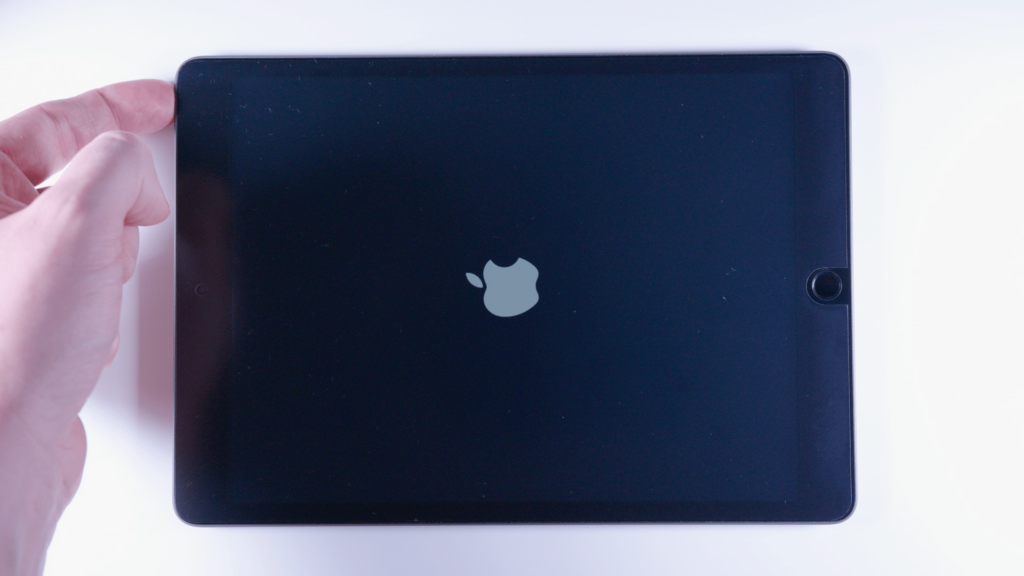 Ein Ipad. Ein Finger tippt auf den Rand links oben. Der Bildschirm ist Schwarz. Man sieht den das Logo von Apple in der Mitte. 
