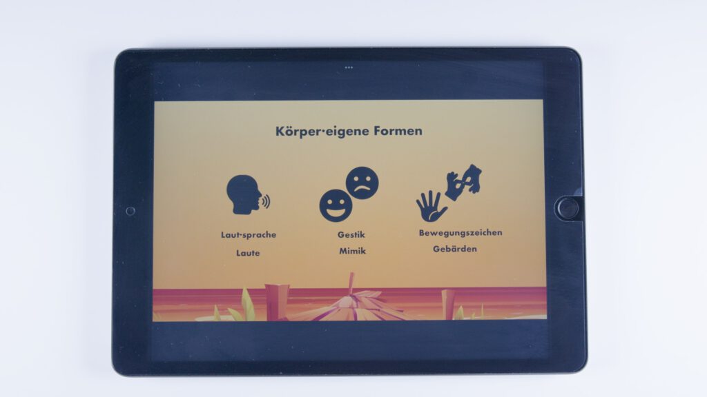 Tablet mit orangem Hintergrund. Überschrift: „Körper·eigene Formen“. Drei Symbole mit Beschriftung: links ein Kopf mit Sprechwellen („Laut·sprache, Laute“), in der Mitte zwei Gesichter mit unterschiedlichen Mimiken („Gestik, Mimik“), rechts Hände in Gebärdenform („Bewegungszeichen, Gebärden“)