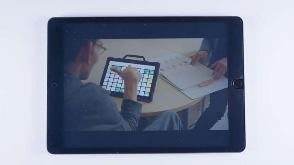 Ein junger Mann nutzt ein Tablet mit Kommunikations-App (Raster mit Symbolfeldern). Er sitzt an einem Tisch, eine zweite Person blättert in einem Ordner mit Piktogrammen.