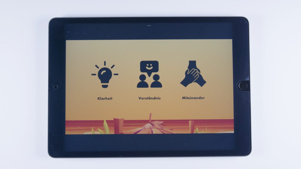 Tablet mit orangem Hintergrund. Drei Symbole: eine Glühbirne mit dem Text „Klarheit“, zwei Personen mit Sprechblase „Verständnis“ und zwei sich haltende Hände mit dem Text „Miteinander“.