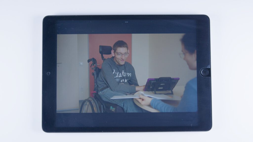 Auf einem Tablet ist ein Foto zu sehen: Ein junger Mann im Rollstuhl nutzt ein Tablet mit Schutzhülle als Kommunikationshilfe. Er sitzt an einem Tisch und lächelt eine Frau an, die ihm ein Dokument zeigt.