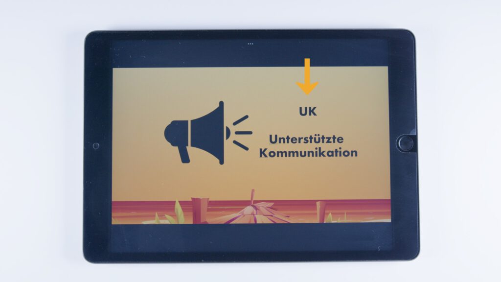 Ein Tablet zeigt auf orangem Hintergrund ein schwarzes Megafon-Symbol mit Schallwellen. Darüber steht in Schwarz: „UK“, darunter: „Unterstützte Kommunikation“. Ein gelber Pfeil zeigt auf die Abkürzung „UK“.
