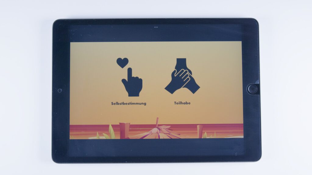 Ein Tablet zeigt auf orangem Hintergrund zwei schwarze Symbole: eine Hand mit Zeigefinger und schwebendem Herz, beschriftet mit „Selbstbestimmung“, und zwei sich haltende Hände, beschriftet mit „Teilhabe“.