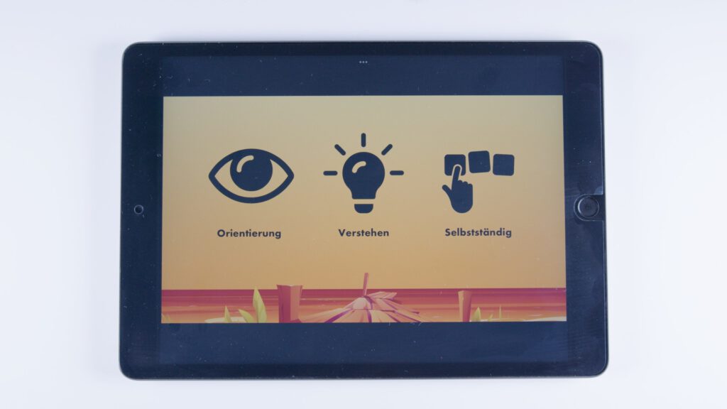 Tablet mit orangem Hintergrund. Drei Symbole: ein Auge mit dem Text „Orientierung“, eine Glühbirne mit dem Text „Verstehen“, eine Hand mit Symbolkarten mit dem Text „Selbstständig“.