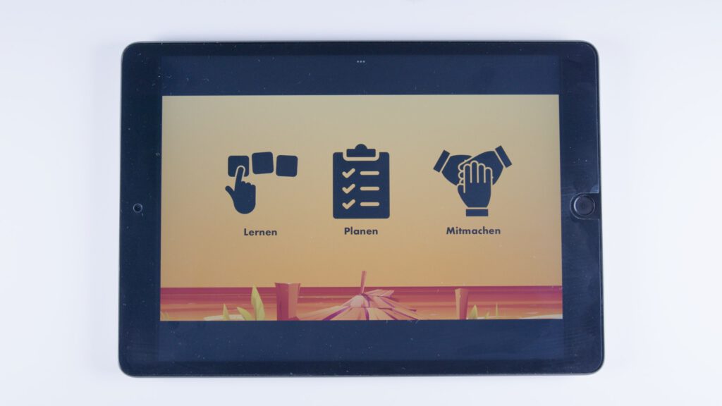 Tablet mit orangem Hintergrund. Drei Symbole: eine Hand mit Symbolkarten (beschriftet mit „Lernen“), eine Checkliste („Planen“) und zwei Hände beim Abklatschen („Mitmachen“).