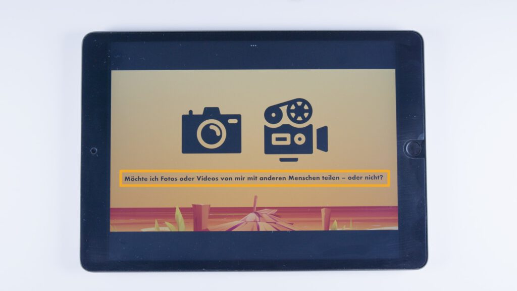 Tablet mit orangem Hintergrund. Zwei Symbole: links eine Kamera, rechts eine Filmkamera. Darunter ein orange hinterlegter Text: „Möchte ich Fotos oder Videos von mir mit anderen Menschen teilen – oder nicht?“.