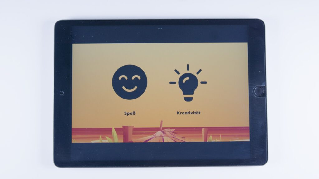 Tablet mit orangem Hintergrund. Links ein lachendes Gesicht mit der Beschriftung „Spaß“, rechts eine Glühbirne mit der Beschriftung „Kreativität“.