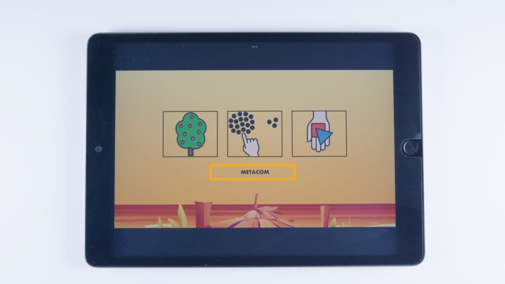 Tablet mit orangem Hintergrund. Drei rechteckige Bildfelder mit METACOM-Symbolen: ein Apfelbaum, eine Hand zeigt auf Punkte, eine Hand hält bunte Karten. Darunter steht „METACOM“.