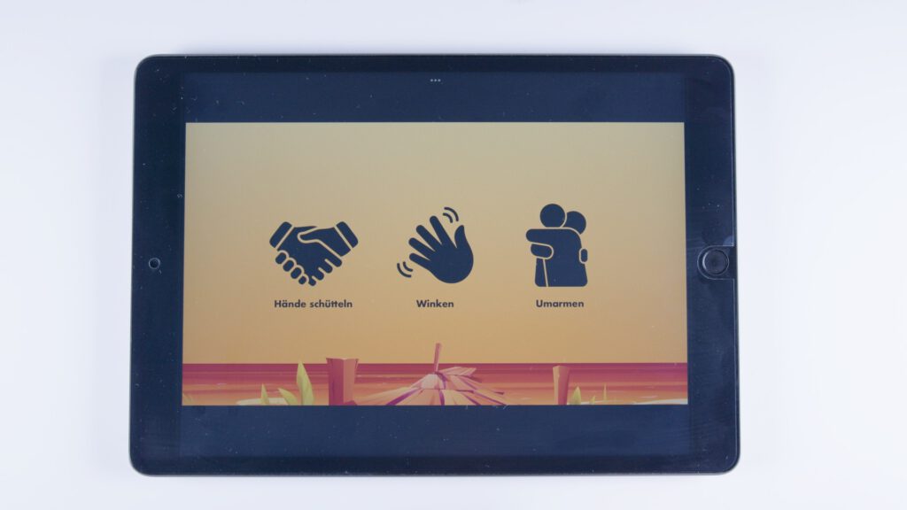 Tablet mit orangem Hintergrund. Drei Symbole: Händeschütteln mit dem Text „Hände schütteln“, winkende Hand mit „Winken“ und zwei umarmende Personen mit der Beschriftung „Umarmen“.