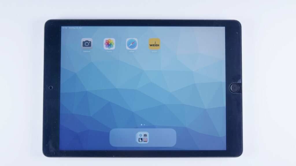 Tablet mit geometrischem Hintergrund. Oben sind die Apps „Kamera“, „Fotos“, „Safari“ und „WEIDI“ in einer Reihe angeordnet. Unten ein App-Ordner mit weiteren Symbolen im Dock.